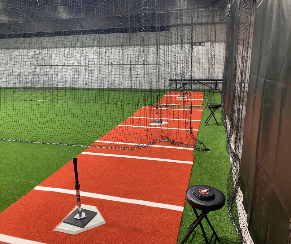 An indoor baseball training facility with a red turf hitting lane marked with white lines, surrounded by black netting. A black tee is set up on the foreground, with additional stands and equipment visible along the lane.